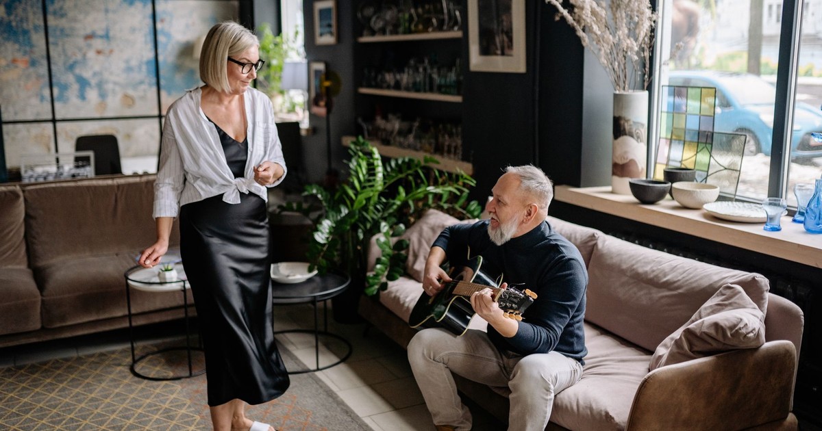 Elderly man playing acoustic guitar while woman listens in cozy living room setting.