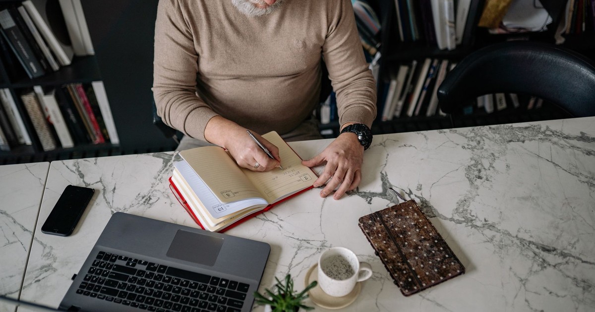 Elderly man writing in a notebook at home office, surrounded by technology and books.