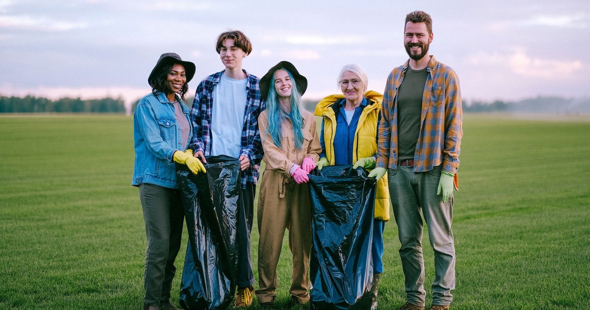 A group of diverse volunteers holding garbage bags while cleaning a green field outdoors.