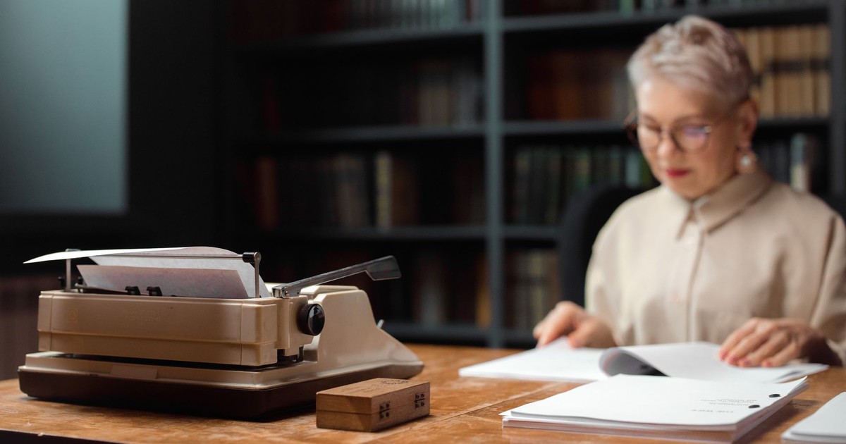 Elderly woman reviewing documents at a desk with a vintage typewriter.