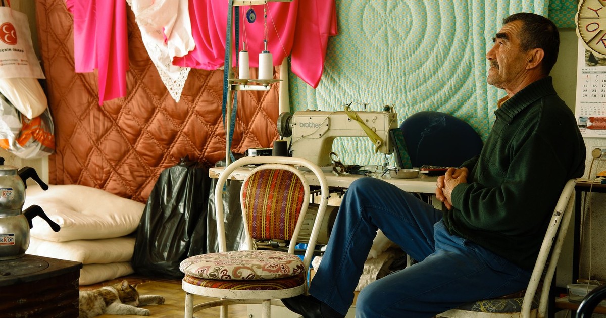 Elderly tailor sitting in his colorful sewing workshop with a cat and sewing machine.
