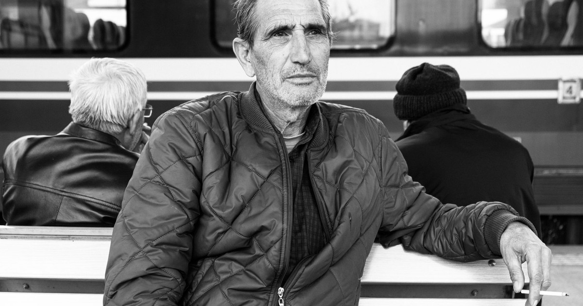 Black and white photo of an elderly man sitting on a bench at a train station, deep in thought.