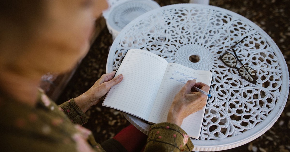Elderly person writing in a notebook at an ornate outdoor table, capturing a peaceful moment.