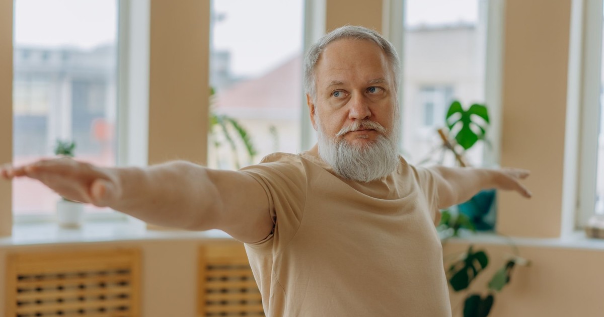 Elderly man performing Warrior Pose yoga for wellness indoors.