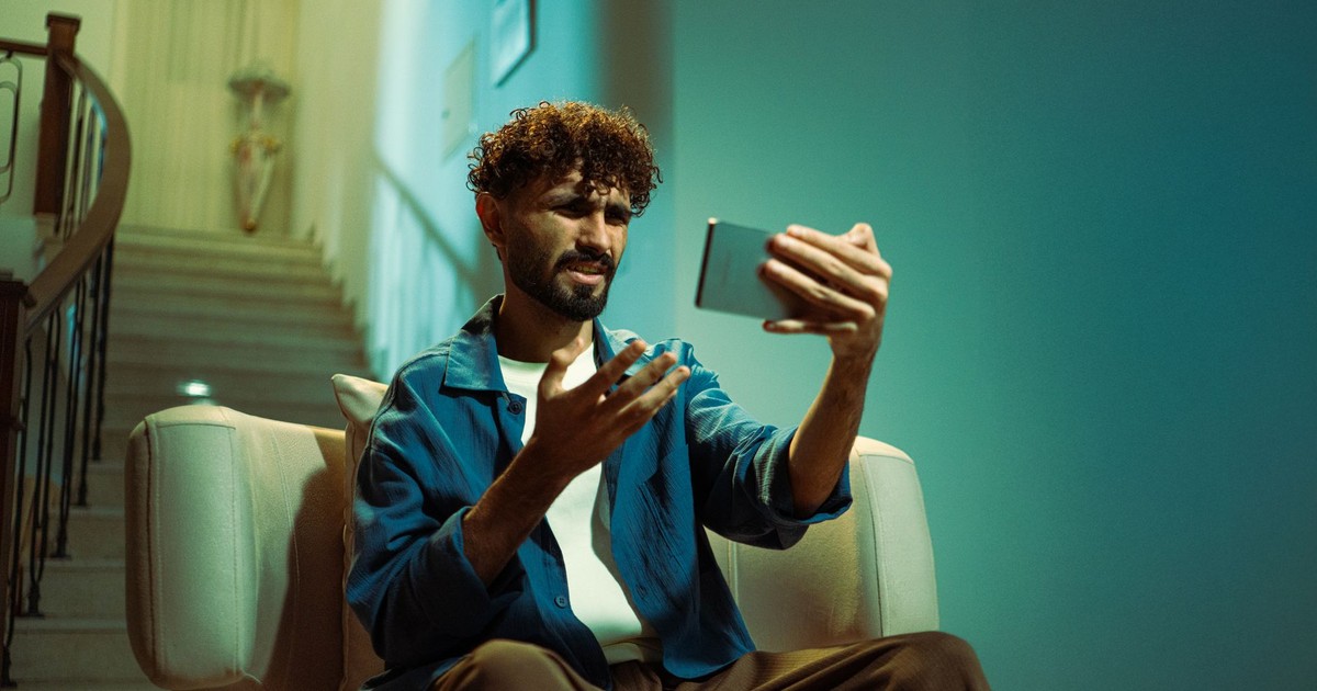 A man sitting on a couch indoors, looking confused while using a smartphone.