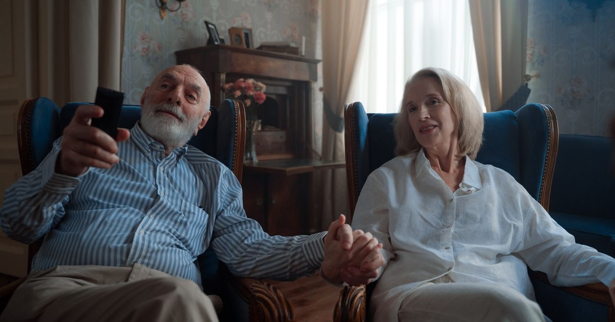 Senior couple sitting together, holding hands and watching TV in a cozy living room.