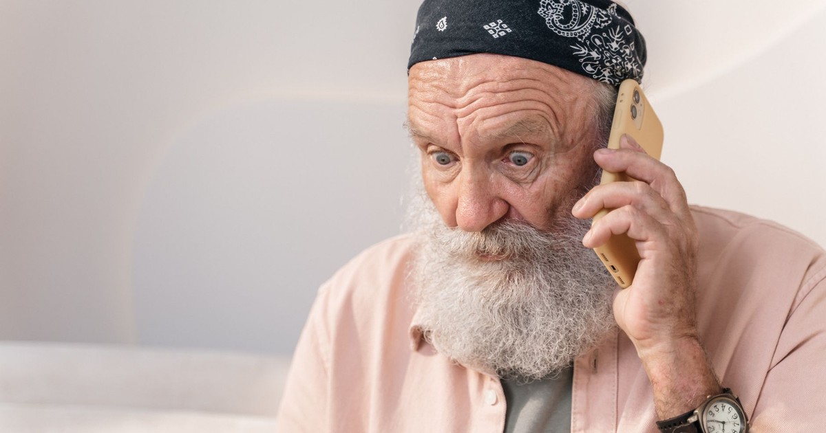 An elderly man with a beard looks surprised while on the phone indoors.