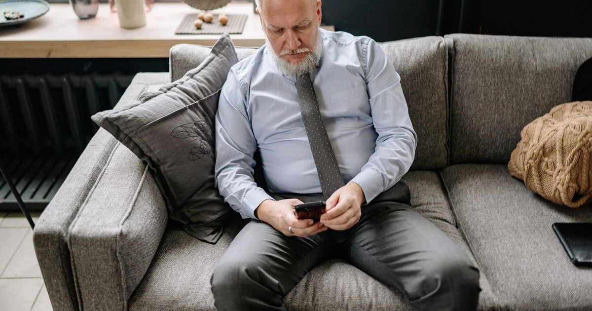 Senior man in formal attire scrolling on phone while sitting on a couch.