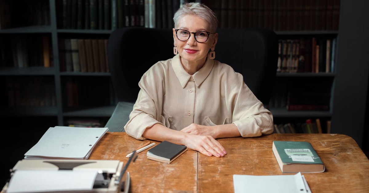 Elderly woman with eyeglasses at a library desk, surrounded by books.