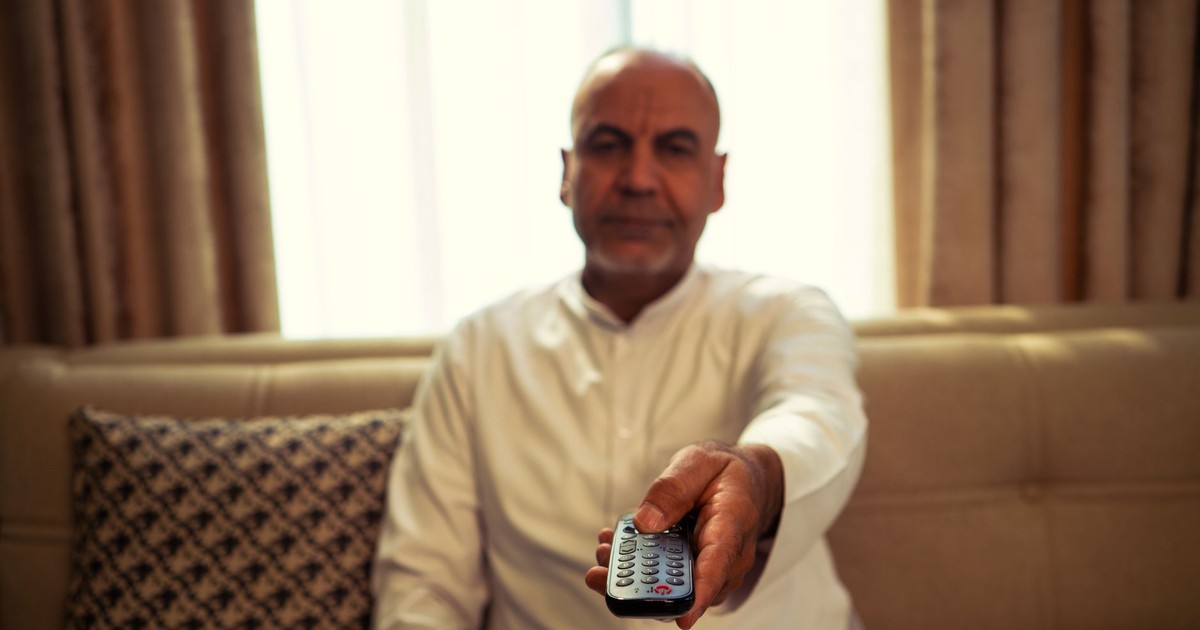 A senior man in a living room extends a TV remote towards the camera.