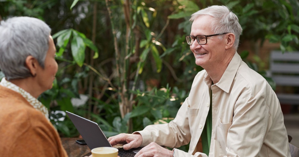 Happy senior couple sitting in a garden cafe, enjoying coffee and conversation with a laptop.