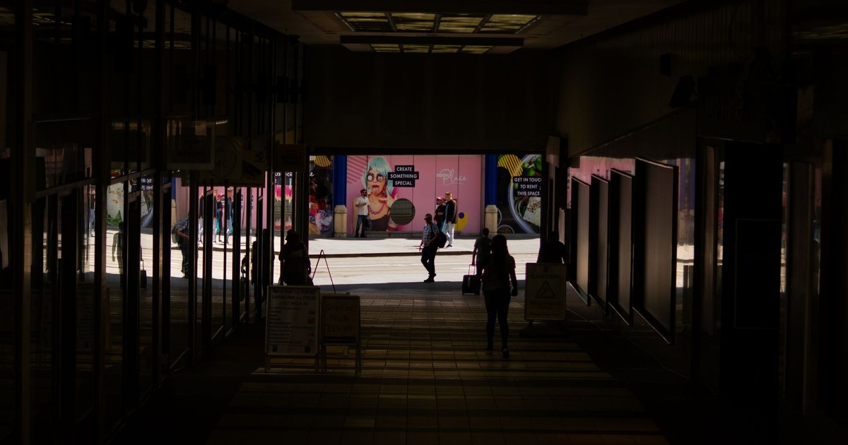 Silhouettes walk through a dimly lit indoor passageway leading to a bright shopping area.