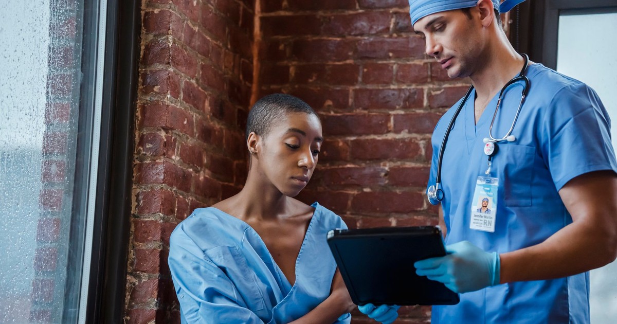 Calm doctor in blue uniform standing with clipboard and demonstrating diagnosis to African American woman in patient robe in hallway of clinic