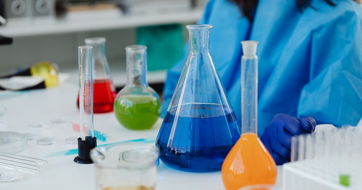 A close-up of a laboratory desk with colorful chemicals in glassware, featuring a researcher in protective gear.