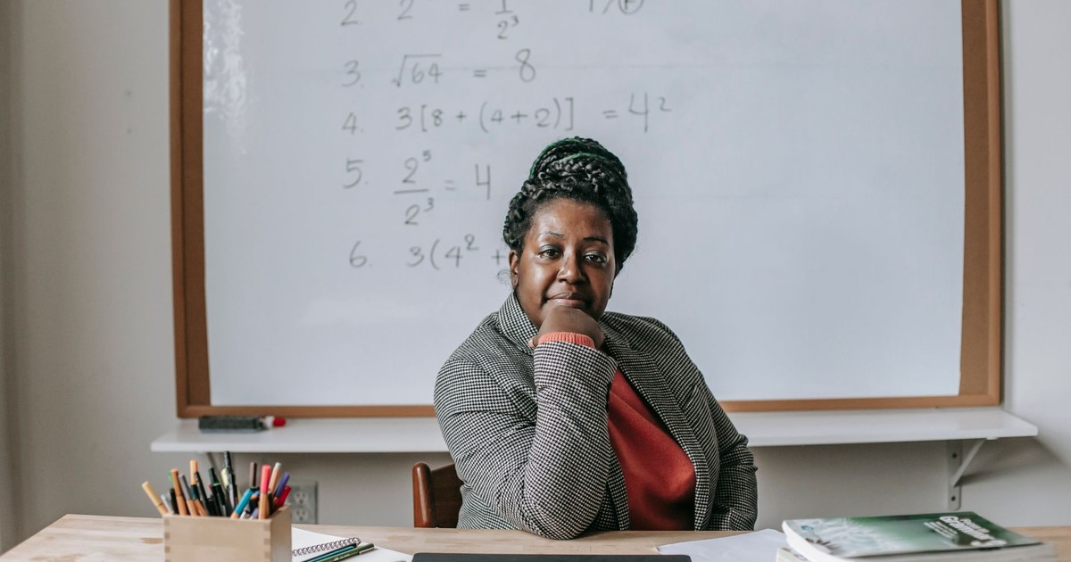 African American female teacher in formal clothes sitting at table with books and laptop and looking at camera