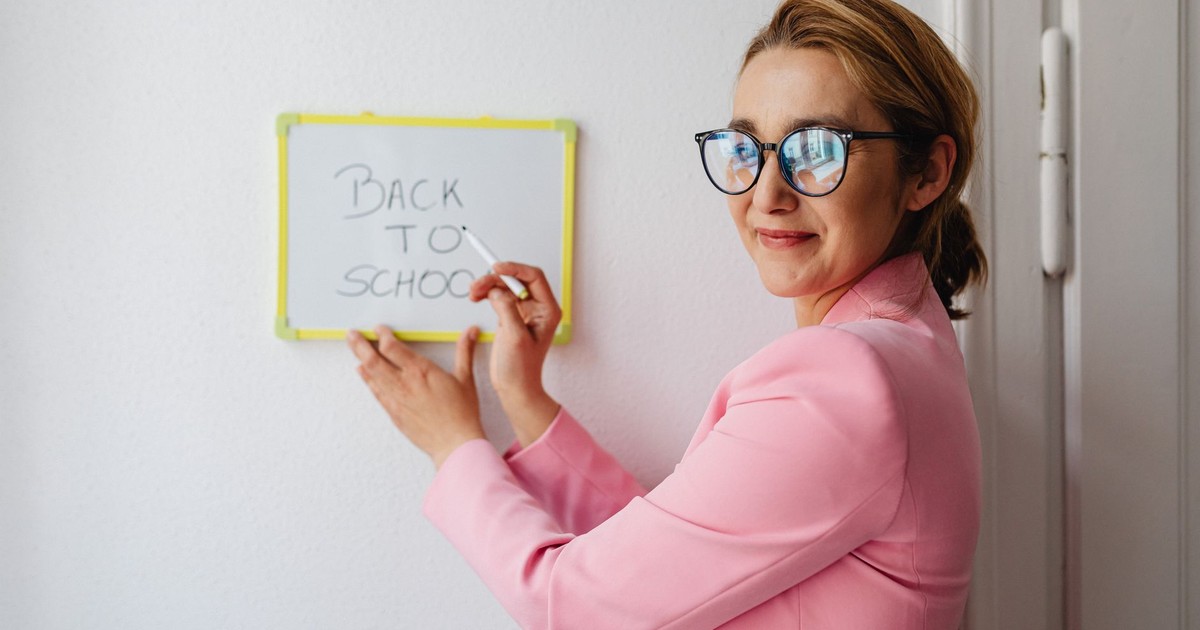 A cheerful female teacher in a pink blazer writes 'Back to School' on a whiteboard.