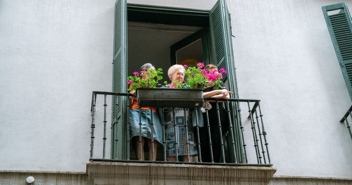 An elderly woman standing on a balcony adorned with vibrant flowers, enjoying the view outdoors.