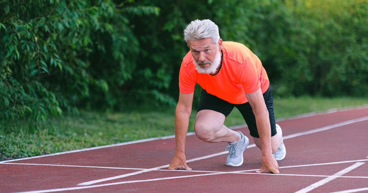 Full body of confident focused senior male ready for sprint on racetrack in stadium near green bushy plants