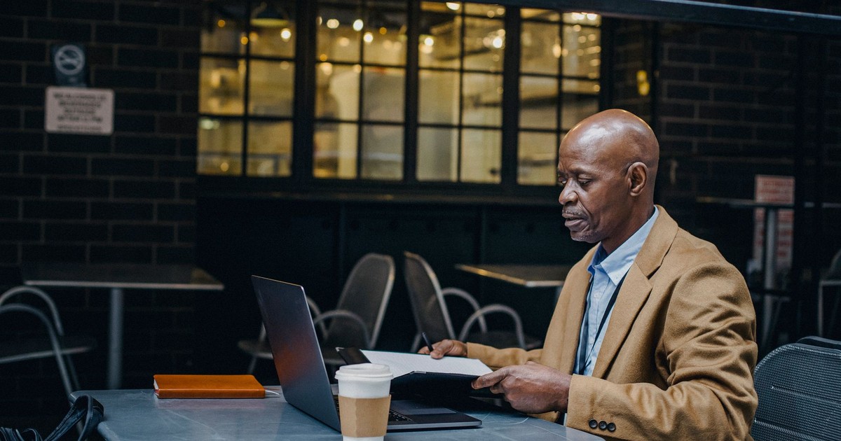 Side view of concentrated African American entrepreneur in stylish clothes using laptop and reading documents in city cafe