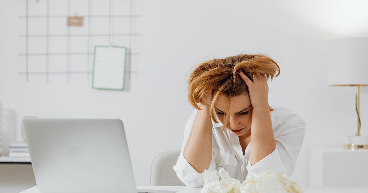 Frustrated woman in white shirt at desk with laptop, feeling overwhelmed.