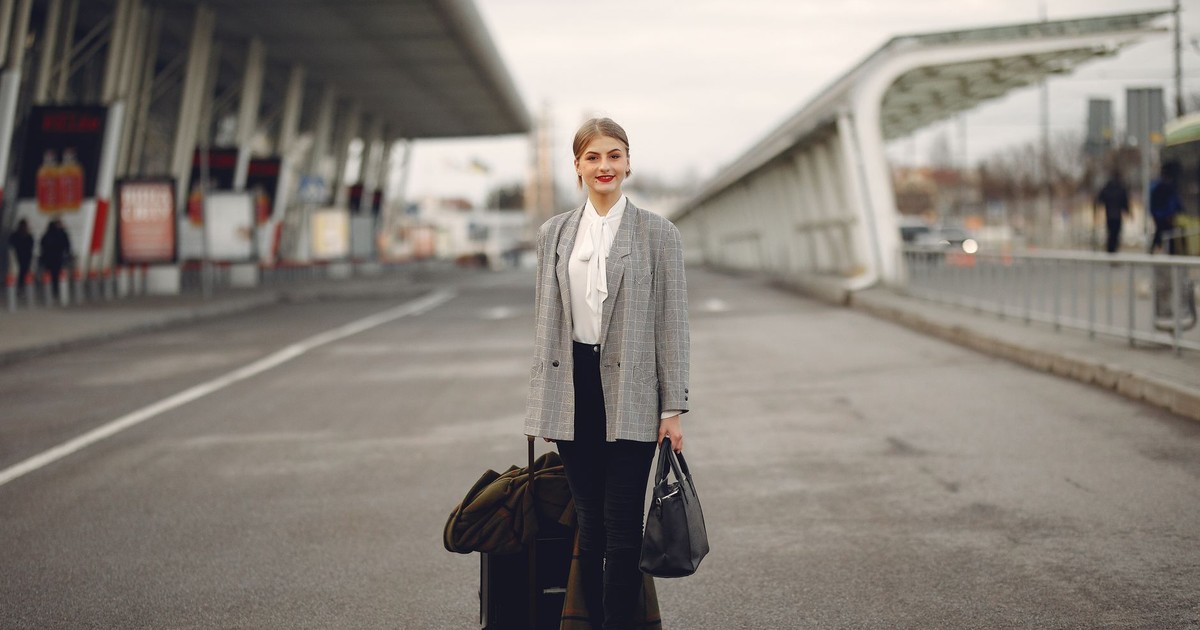 Full body of young woman in stylish outfit carrying suitcase and bag while walking along pavement near modern airport building
