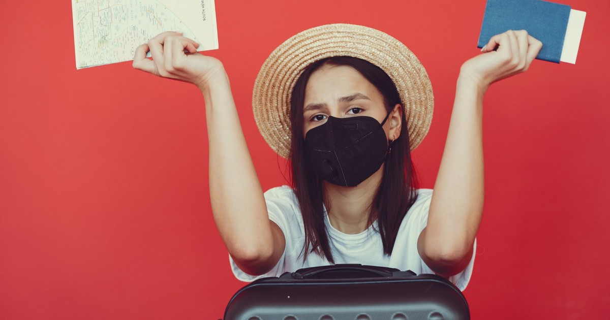 Upset young lady wearing straw hat and protective mask with map and passport leaning on black suitcase and looking at camera with disappointment on red background
