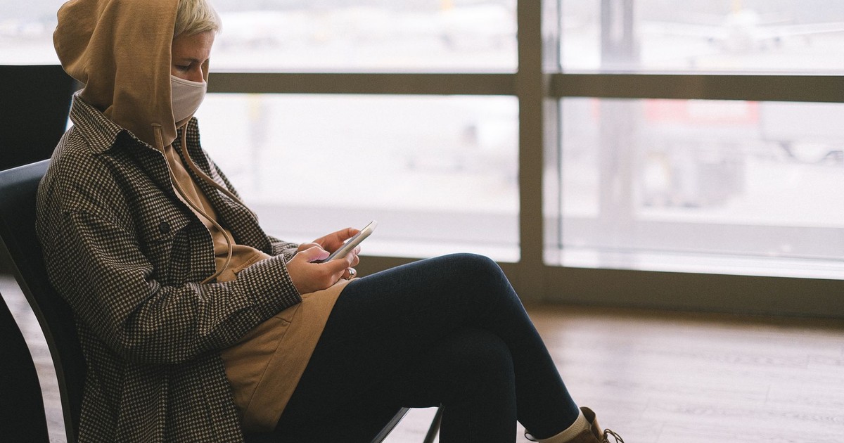 A woman in a hoodie and face mask uses a smartphone while waiting at an airport terminal.