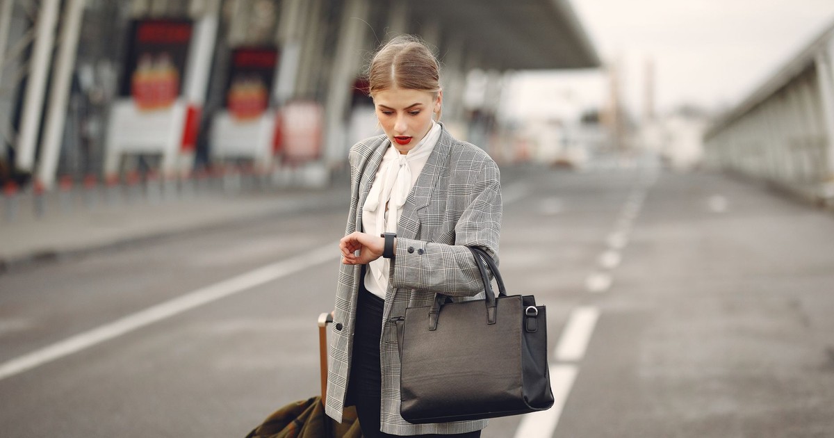 Pensive female passenger in formal outfit checking time on wristwatch while walking along asphalt road to airport terminal with suitcase and handbag during business trip