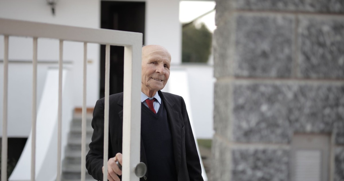 An elderly man in a suit joyfully opens the gate to his home, enjoying the daylight.