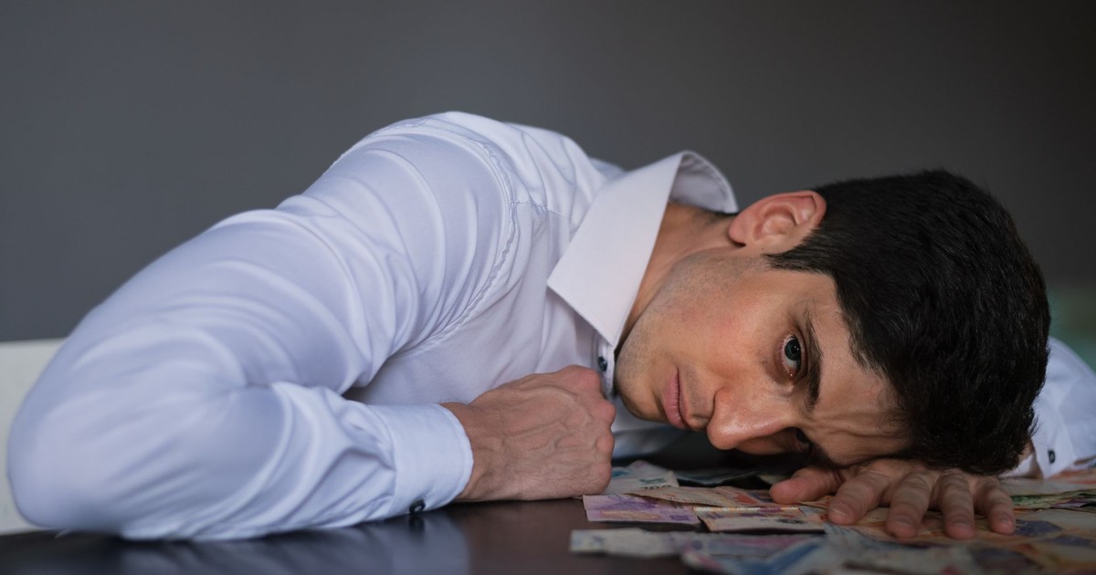 Adult man in shirt lying on table, surrounded by currency, looking stressed indoors.