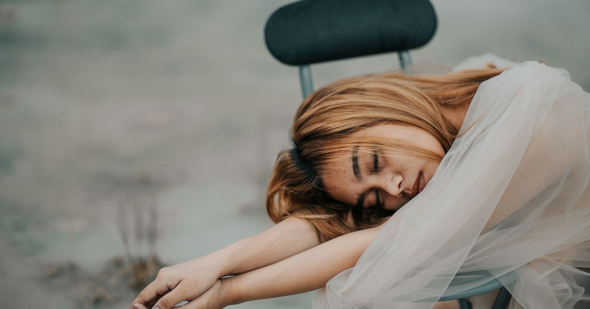 Young dreamy ethnic female with dyed hair and closed eyes covered with transparent fabric on chair