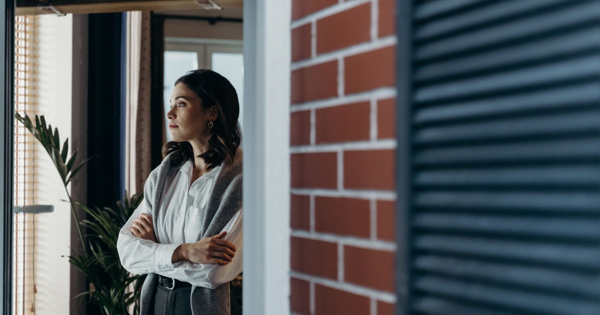 A contemplative woman stands indoors, arms crossed, gazing out the window beside a brick wall.