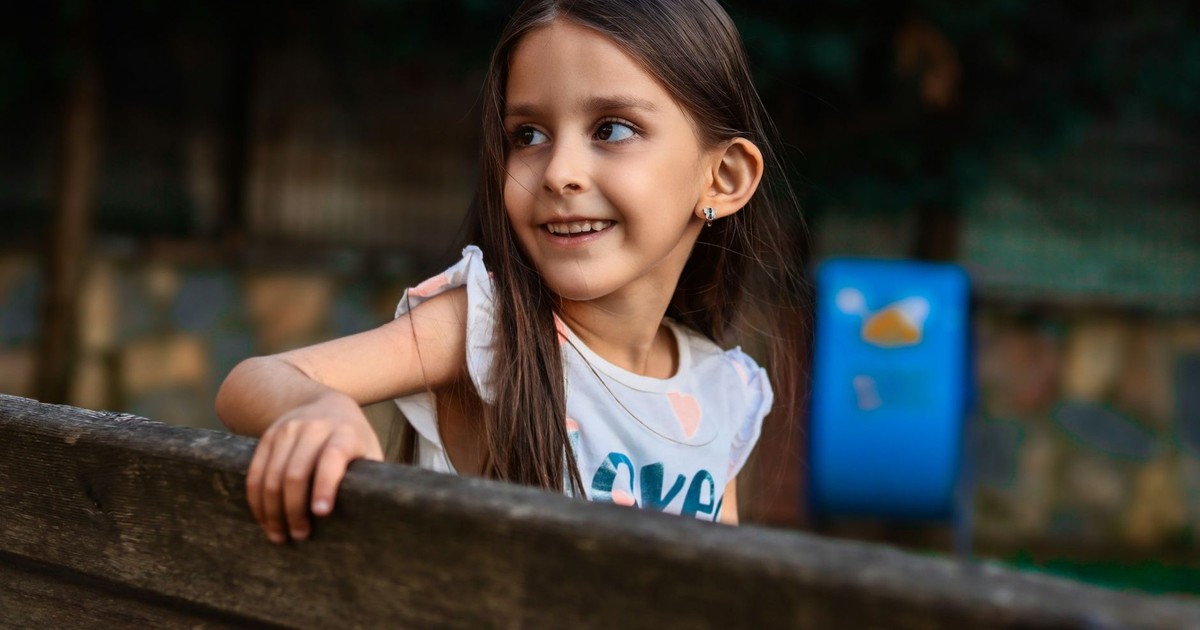 Young girl smiling while leaning on a wooden bench outdoors in a park setting.