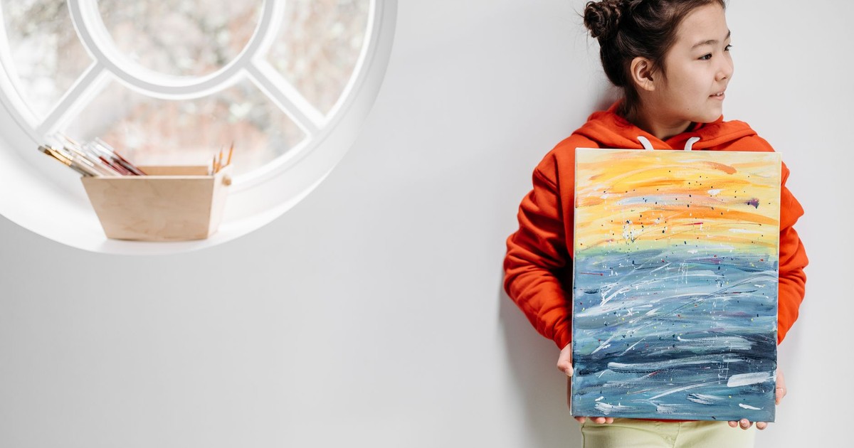 A young girl in a bright hoodie proudly holding her colorful painting indoors.