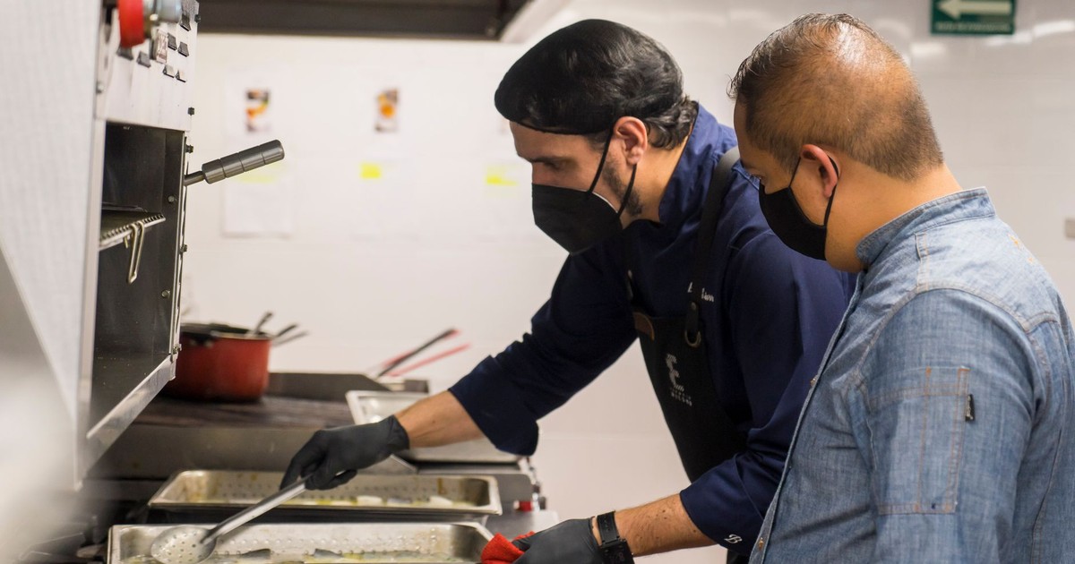 Two chefs in masks and gloves cooking in a professional kitchen setting.