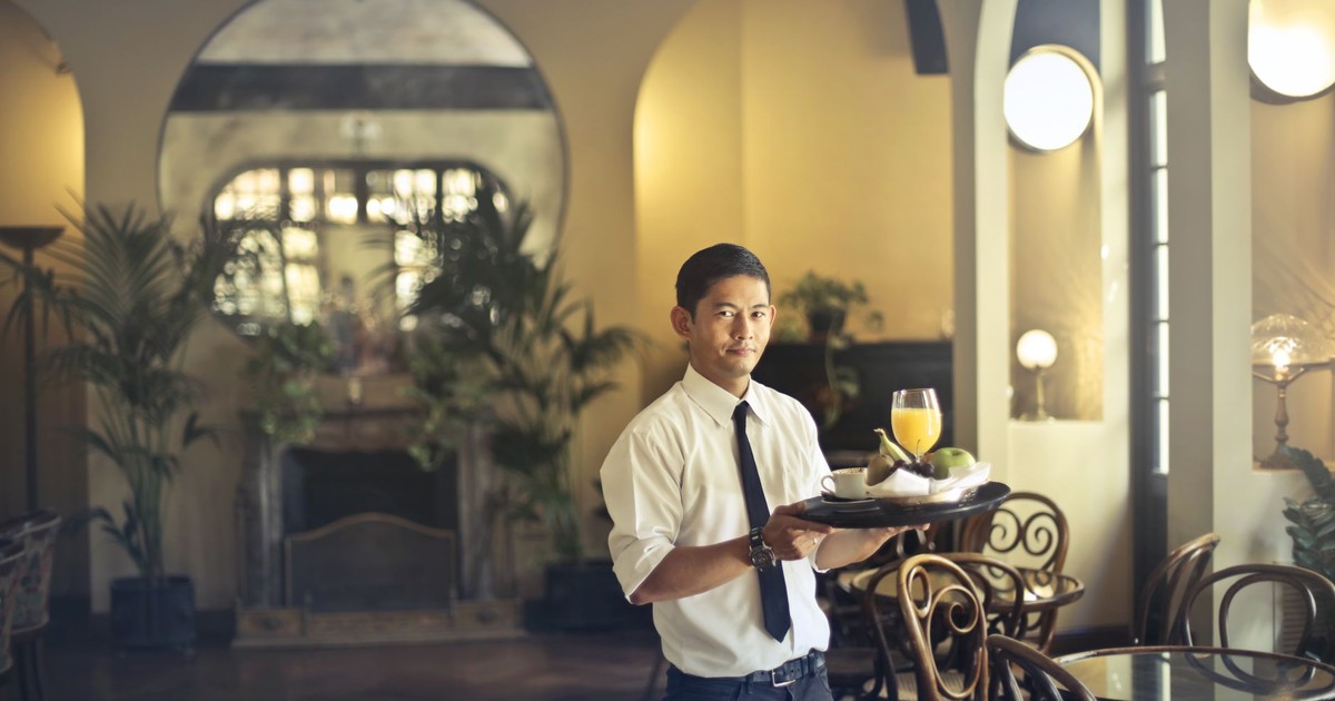 Confident young ethnic waiter in elegant clothes holding tray with food and drinks and looking at camera while serving tables in stylish restaurant