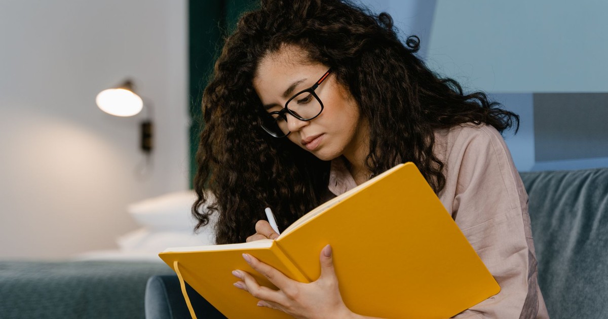 Young woman with curly hair writes in a yellow notebook, seated indoors.