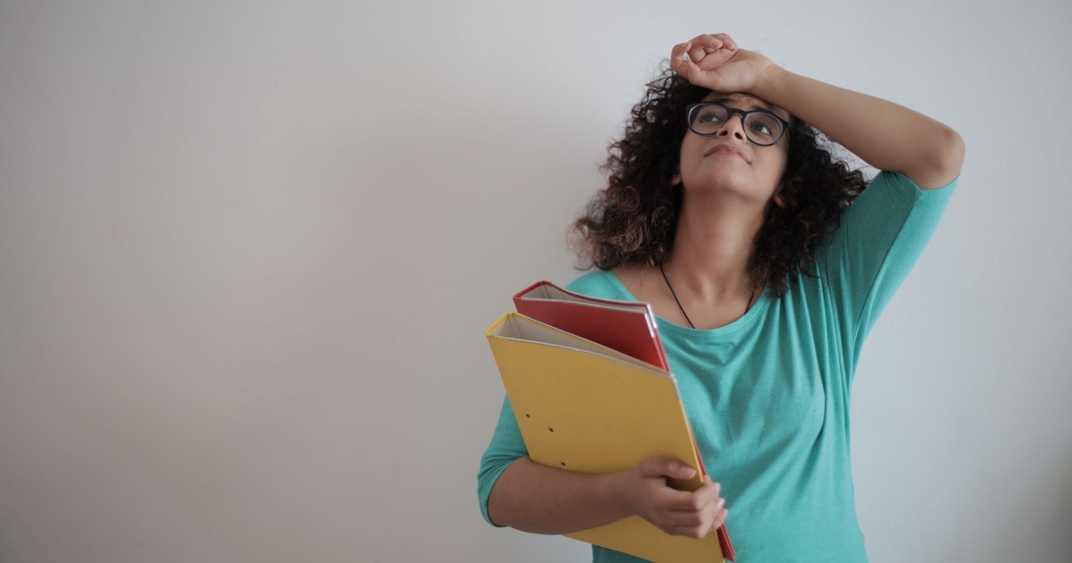 Low angle of unhappy exhausted woman in glasses and casual clothes with folders of papers looking up with sorrow and touching forehead while standing against white wall and having headache due to workload in office