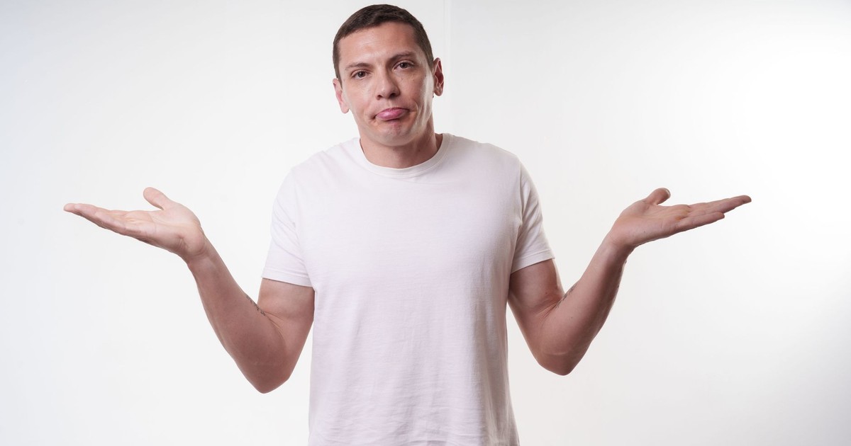 Adult man in white t-shirt shrugs in studio, expressing confusion against a plain background.