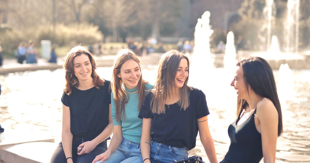 Four friends laughing and chatting outdoors by a water fountain on a sunny day.