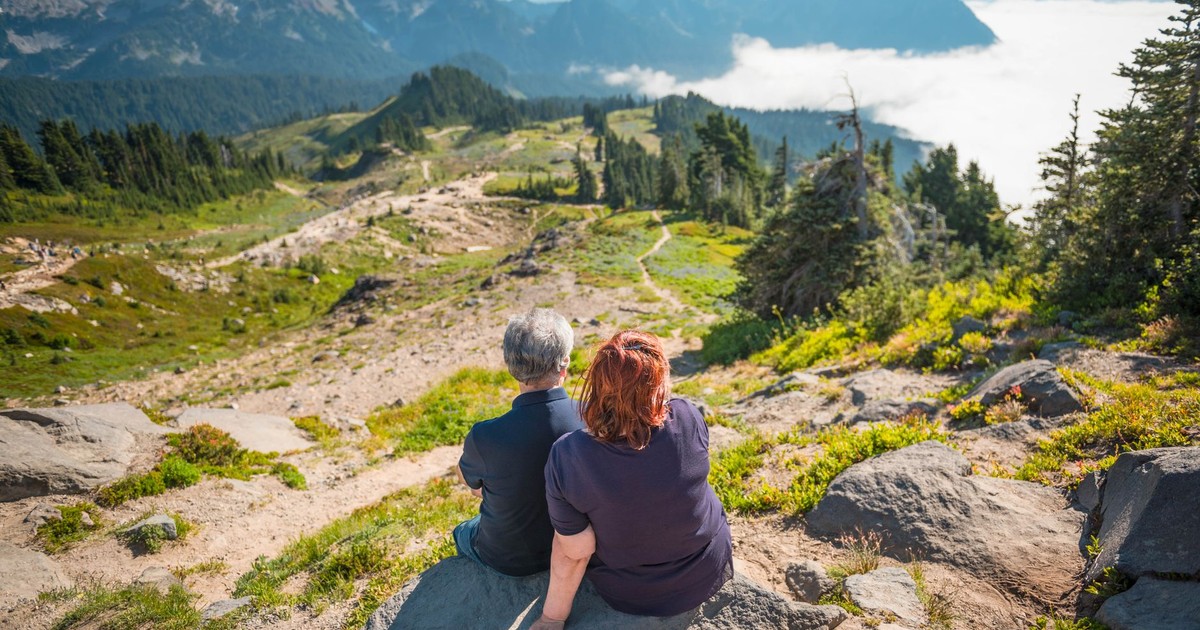 Two senior adults sitting on rocks, admiring a stunning mountain landscape.