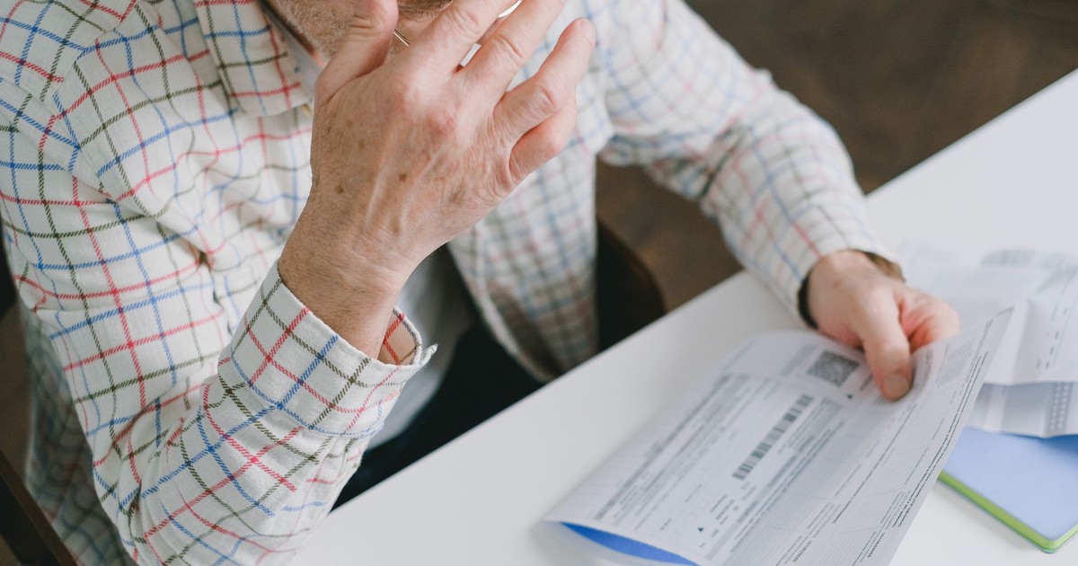 Elderly man in patterned shirt reading and holding bills at a home table, appearing focused.
