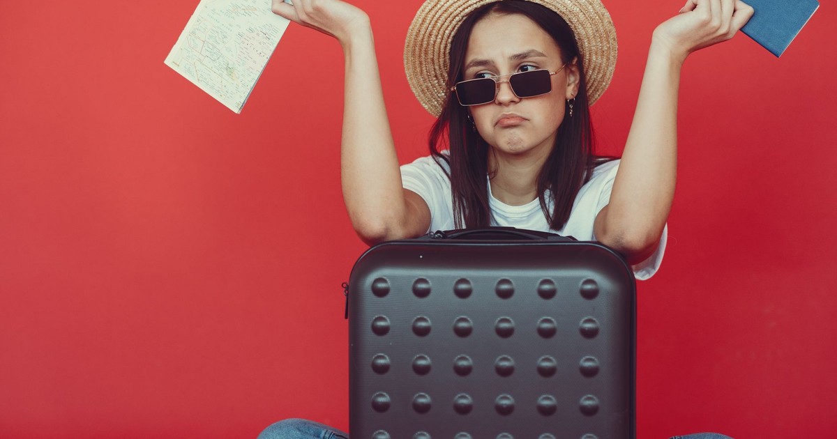 Doubters young stylish woman in wicker hat and trendy sunglasses sitting behind black suitcase and shrugging with passport and tickets on red background