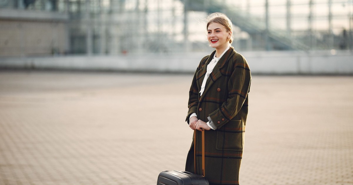 Side view of cheerful stylish blond woman in casual wear with big suitcase standing against airport and waiting for flight