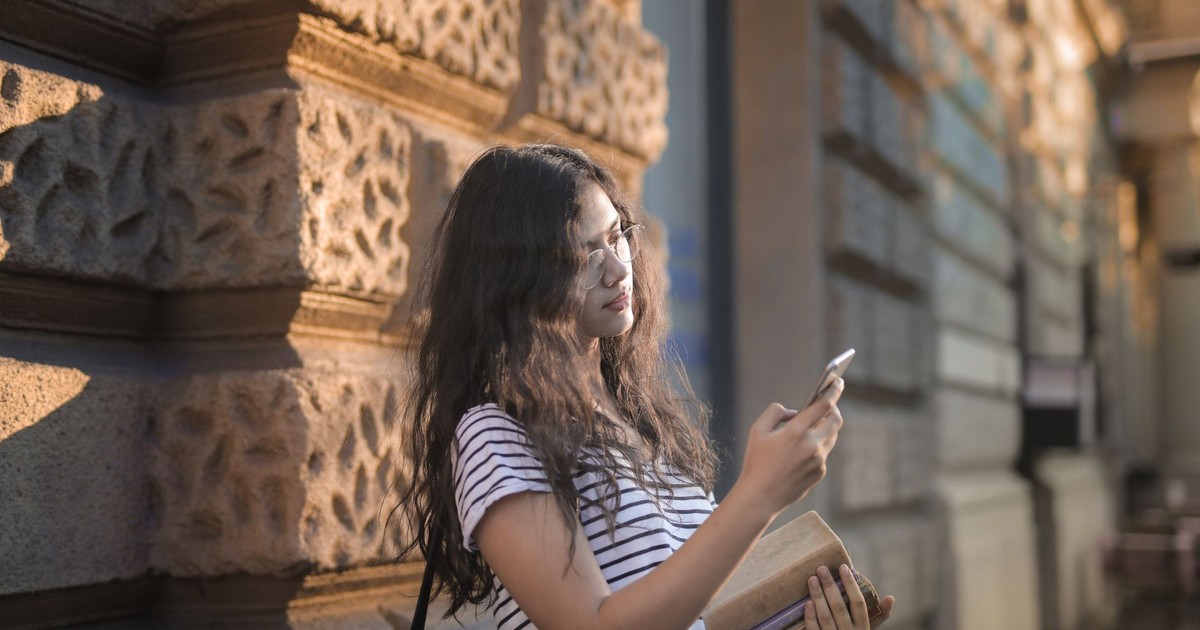 Casual portrait of a young woman leaning on a wall, using her smartphone and holding books outdoors.