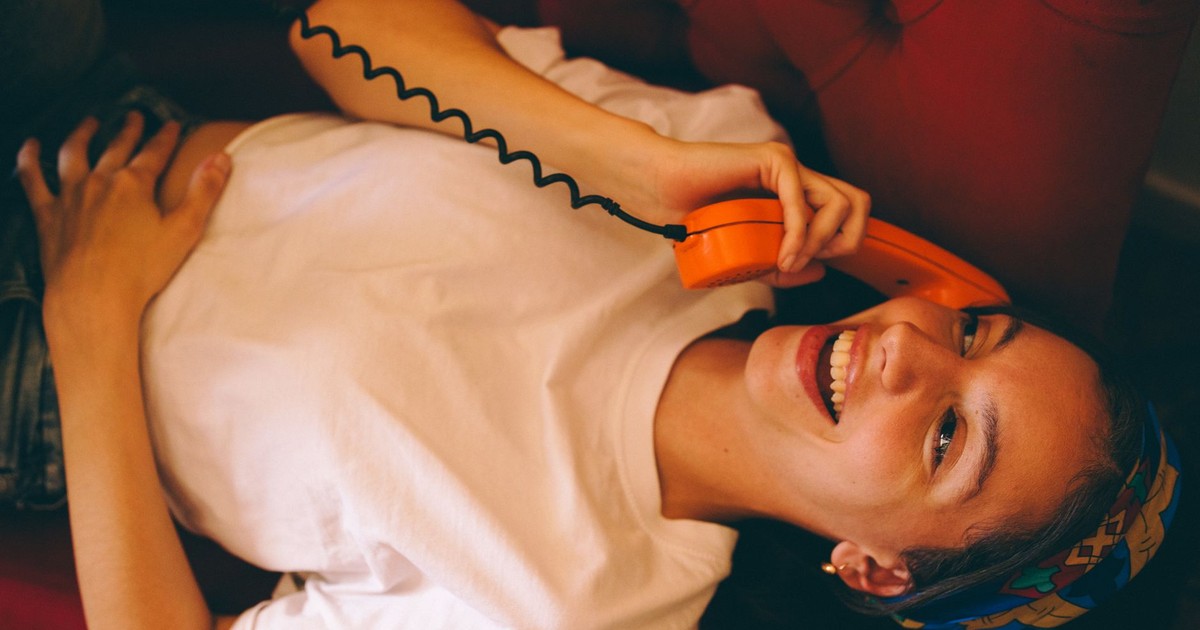 A smiling woman lying on a sofa holds a vintage orange telephone, indoors.