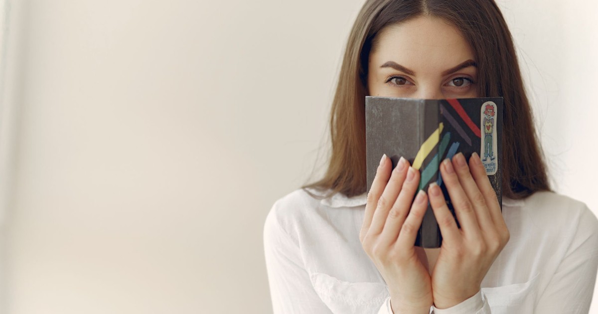Happy young woman with long brown hair in casual clothes looking at camera and covering face with notepad while hiding astonished