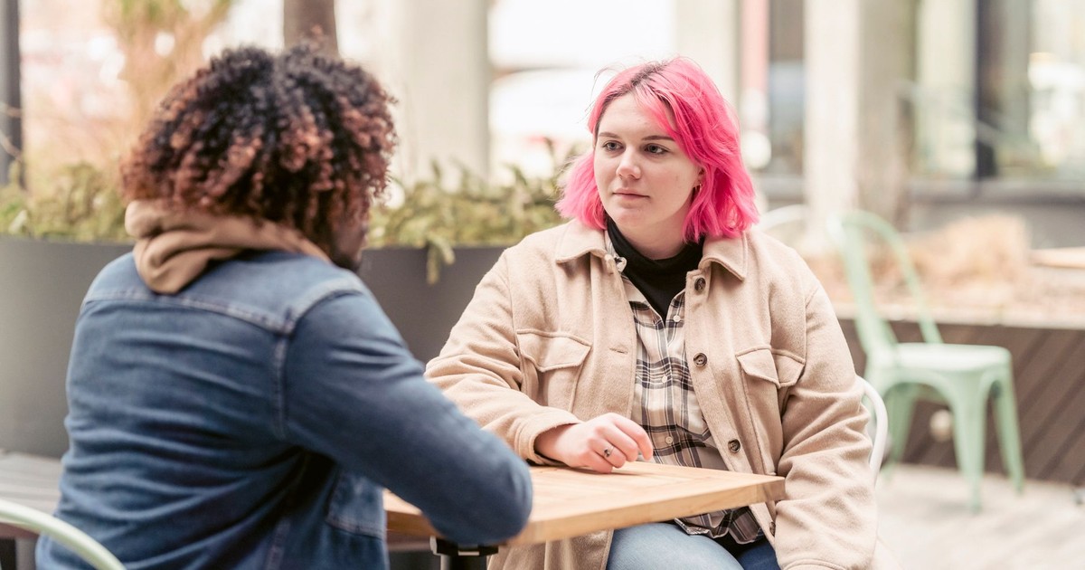 Young woman with pink hair listening to black man while sitting at table in weekend in outdoor cafeteria