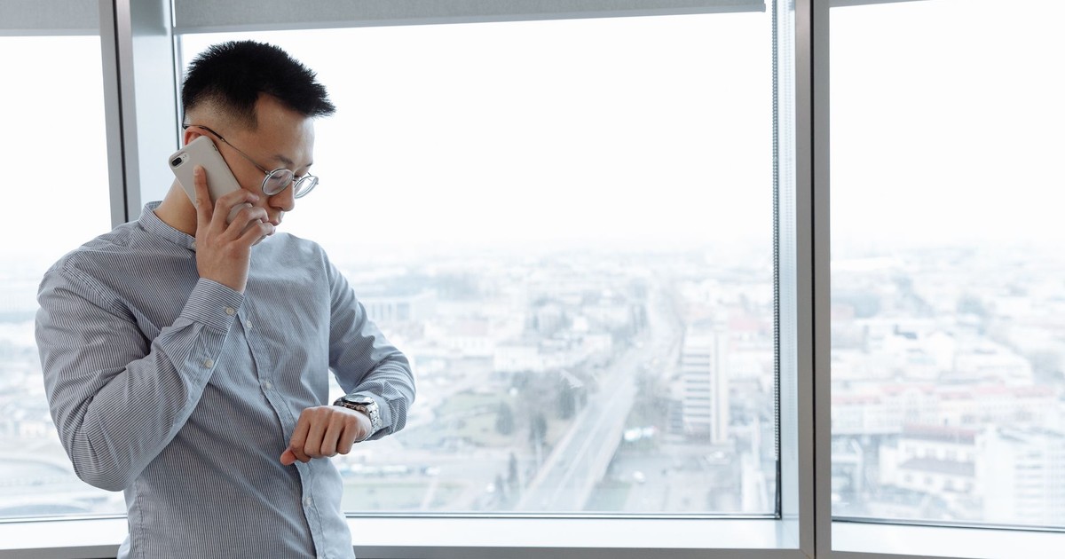 Businessman on a phone call inside a modern office, checking his watch, with a city view.
