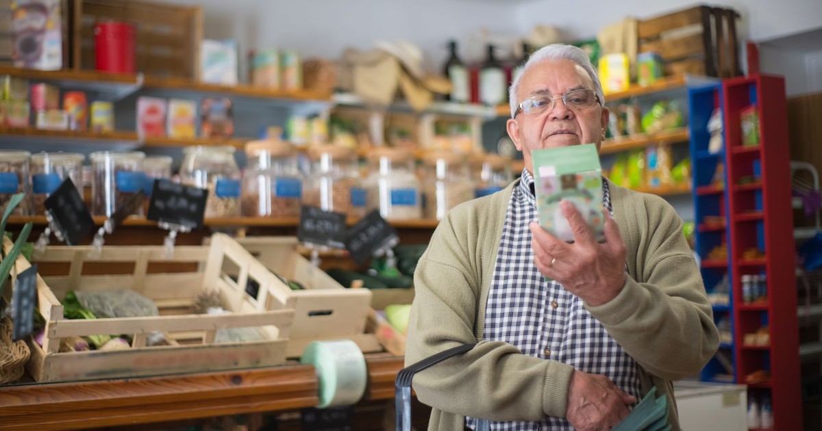 Elderly man examines product in grocery store aisle, representing daily shopping routine.